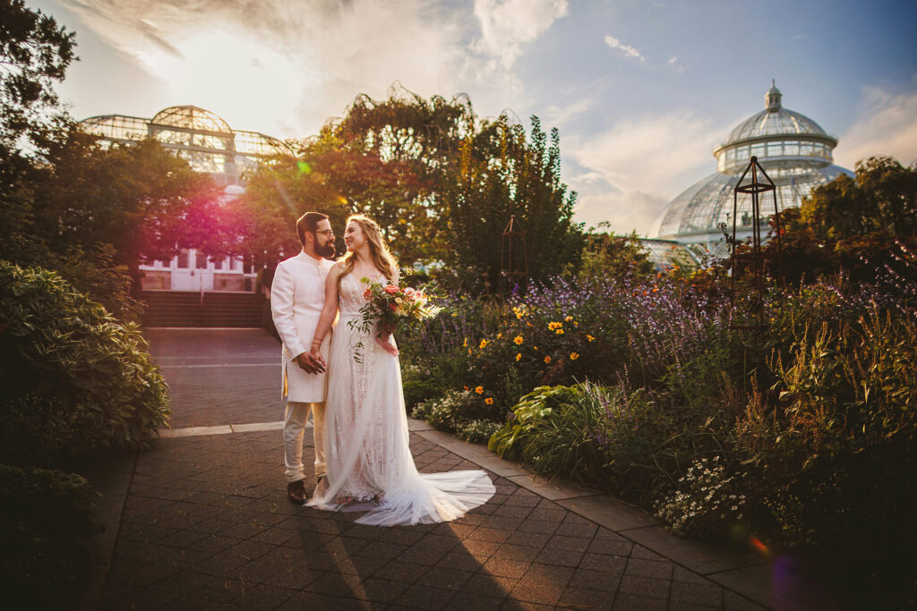 Bride and groom cuddling in front of a large green house backlit by the setting sun